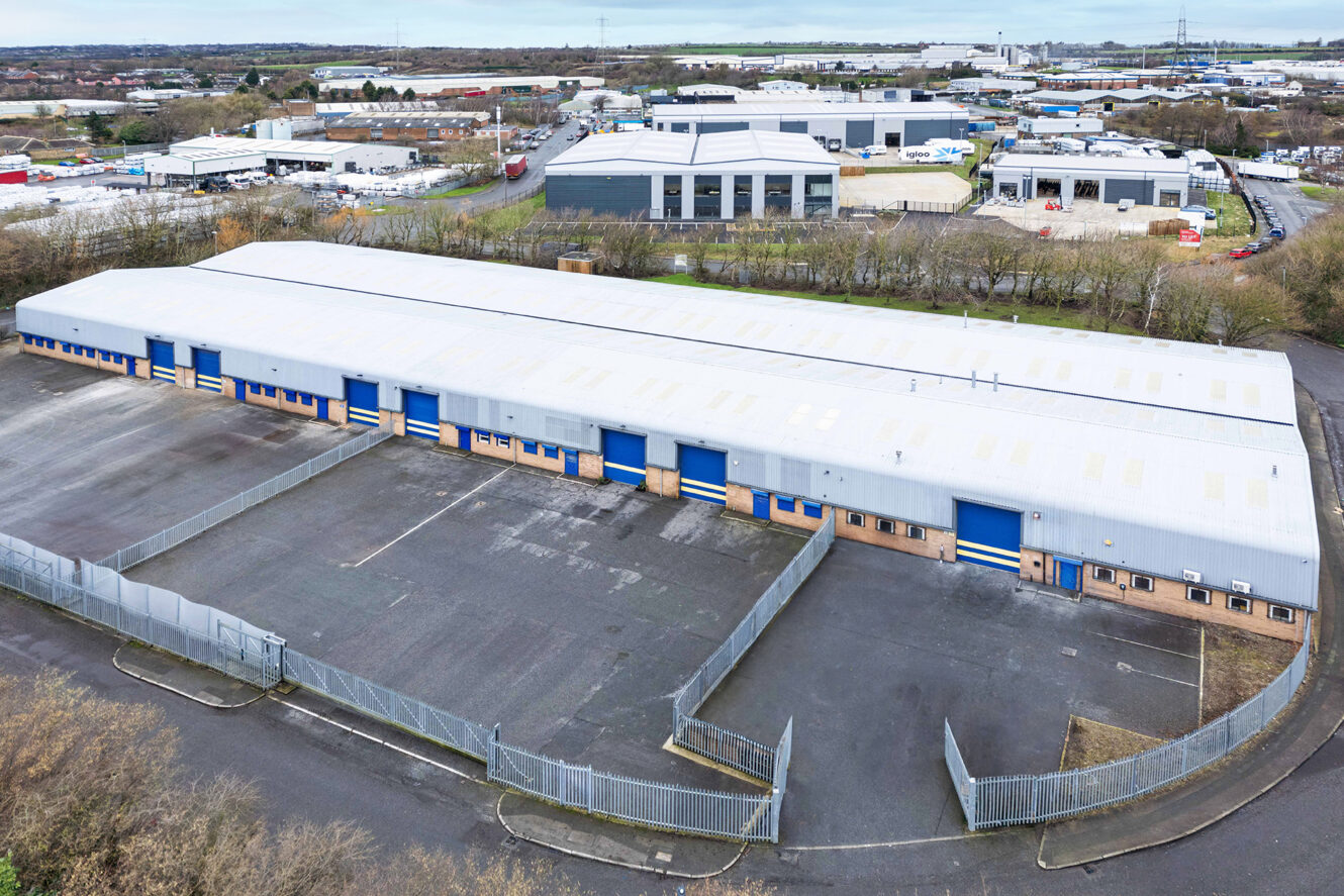 A large industrial warehouse with multiple blue doors, surrounded by fencing, located in a commercial area with other industrial buildings.