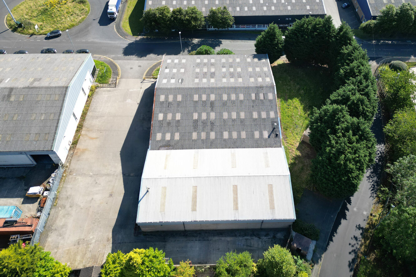 Aerial view of two adjacent industrial warehouses surrounded by trees, with paved lots and nearby roads visible.