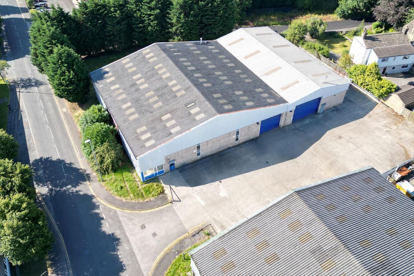 Aerial view of a large industrial warehouse with blue doors, surrounded by paved areas, trees, and nearby roads.