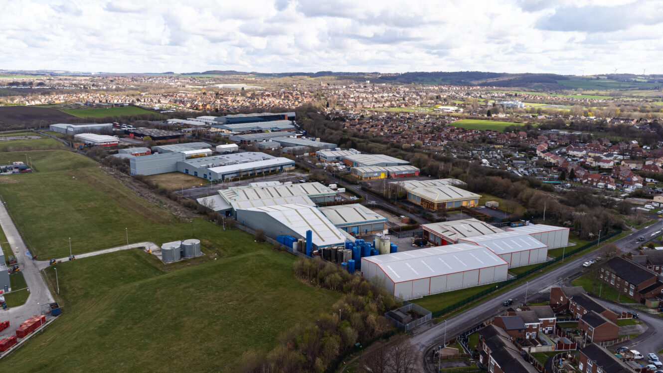 Aerial view of an industrial estate with large warehouses and factory buildings, surrounded by residential neighborhoods and green fields under a cloudy sky.