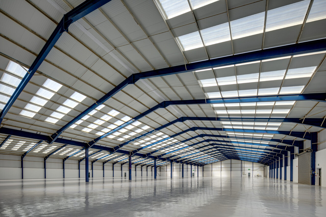 Large, empty industrial warehouse with high ceilings, blue steel beams, and rows of skylights letting in natural light. The polished floor reflects the interior structure.