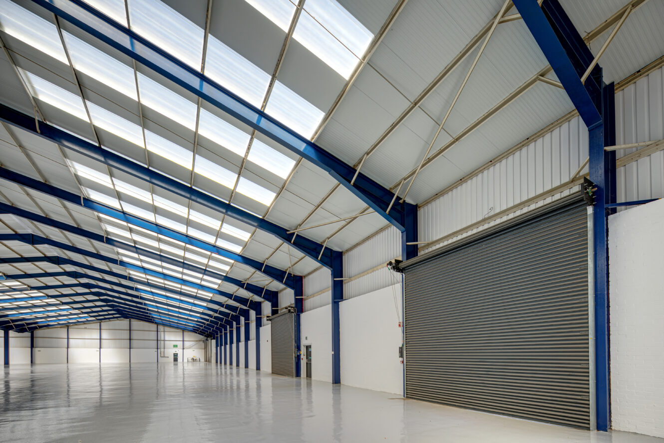 Interior of a large empty warehouse with a high ceiling, blue structural beams, skylights, white walls, and a closed gray roller shutter door.