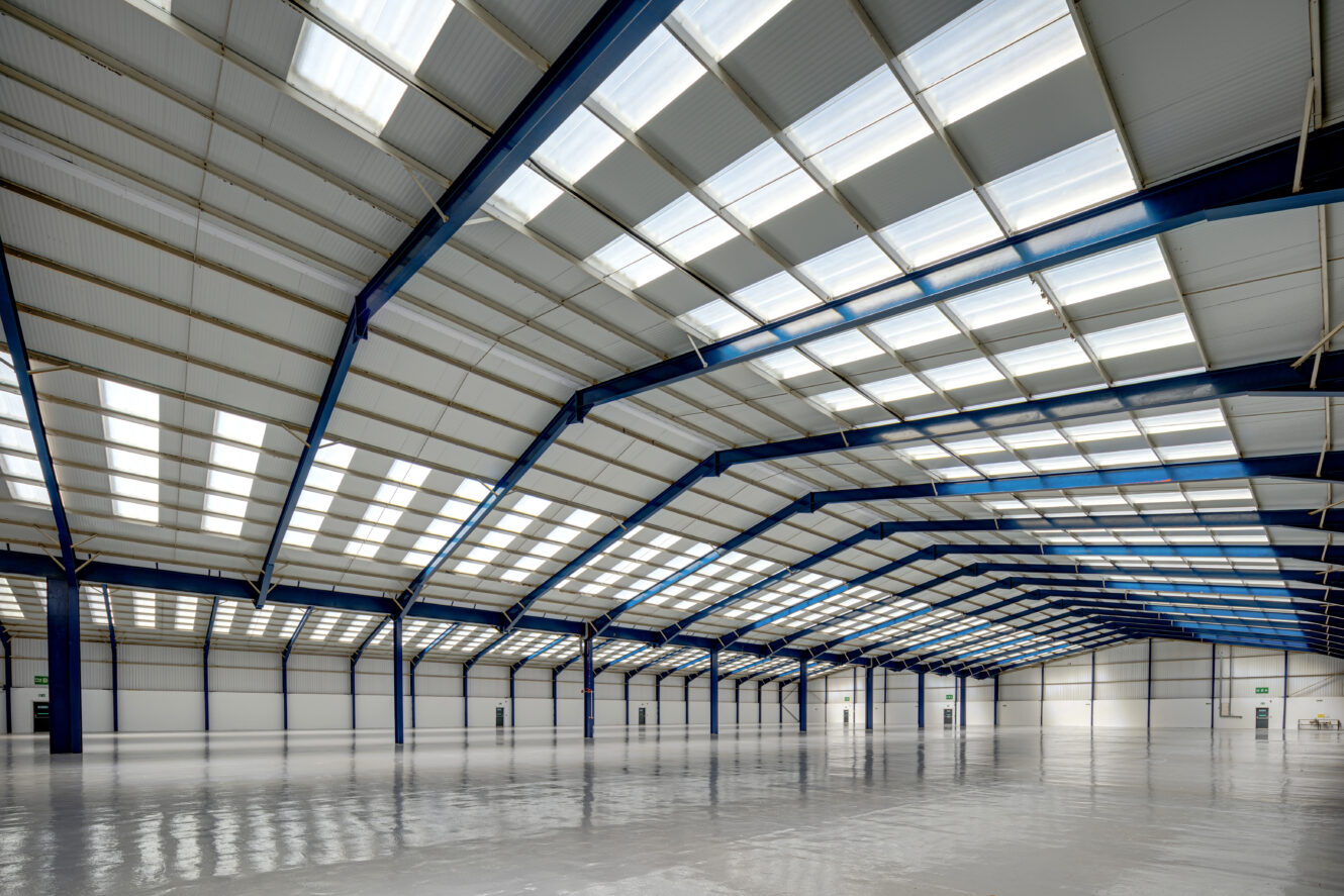 Large, empty warehouse interior with blue steel beams, white walls, and a high, partially skylit ceiling. The polished floor reflects the overhead light.