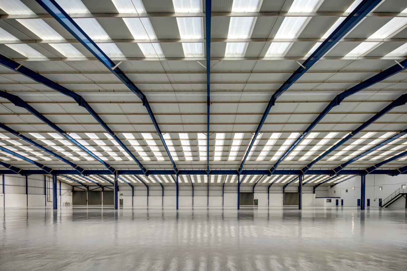 Empty industrial warehouse with high ceilings, steel beams, polished concrete floor, and bright overhead lighting. Walls are white with several doors and a staircase visible.