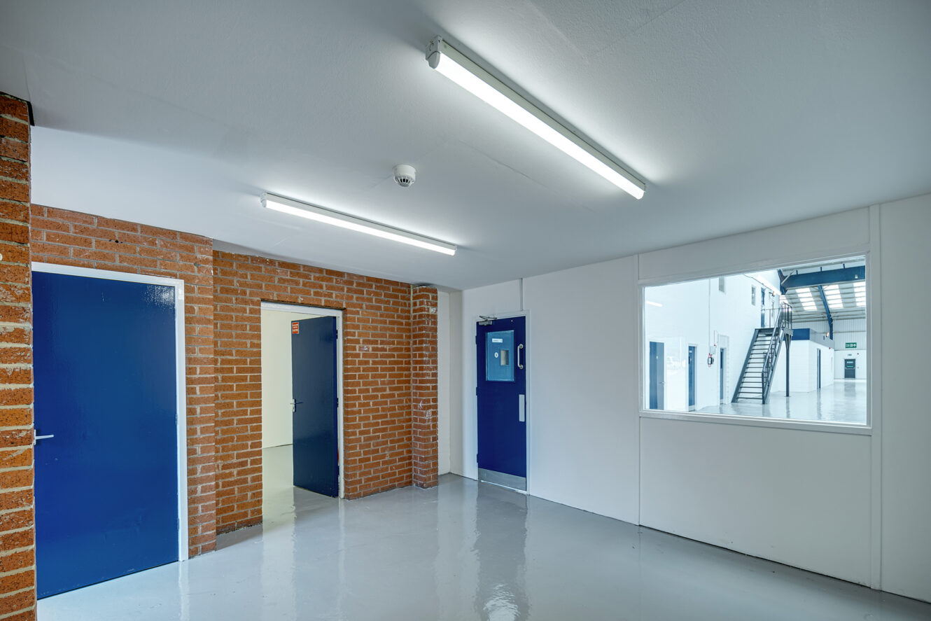 A clean hallway with brick and white walls, blue doors, fluorescent lighting, and a window looking into another corridor with stairs.