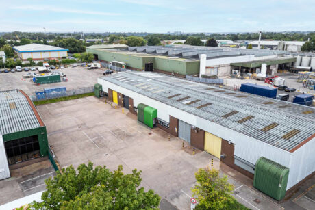Aerial view of an industrial complex with large warehouses, loading bays, parked vehicles, and open paved areas under a cloudy sky.