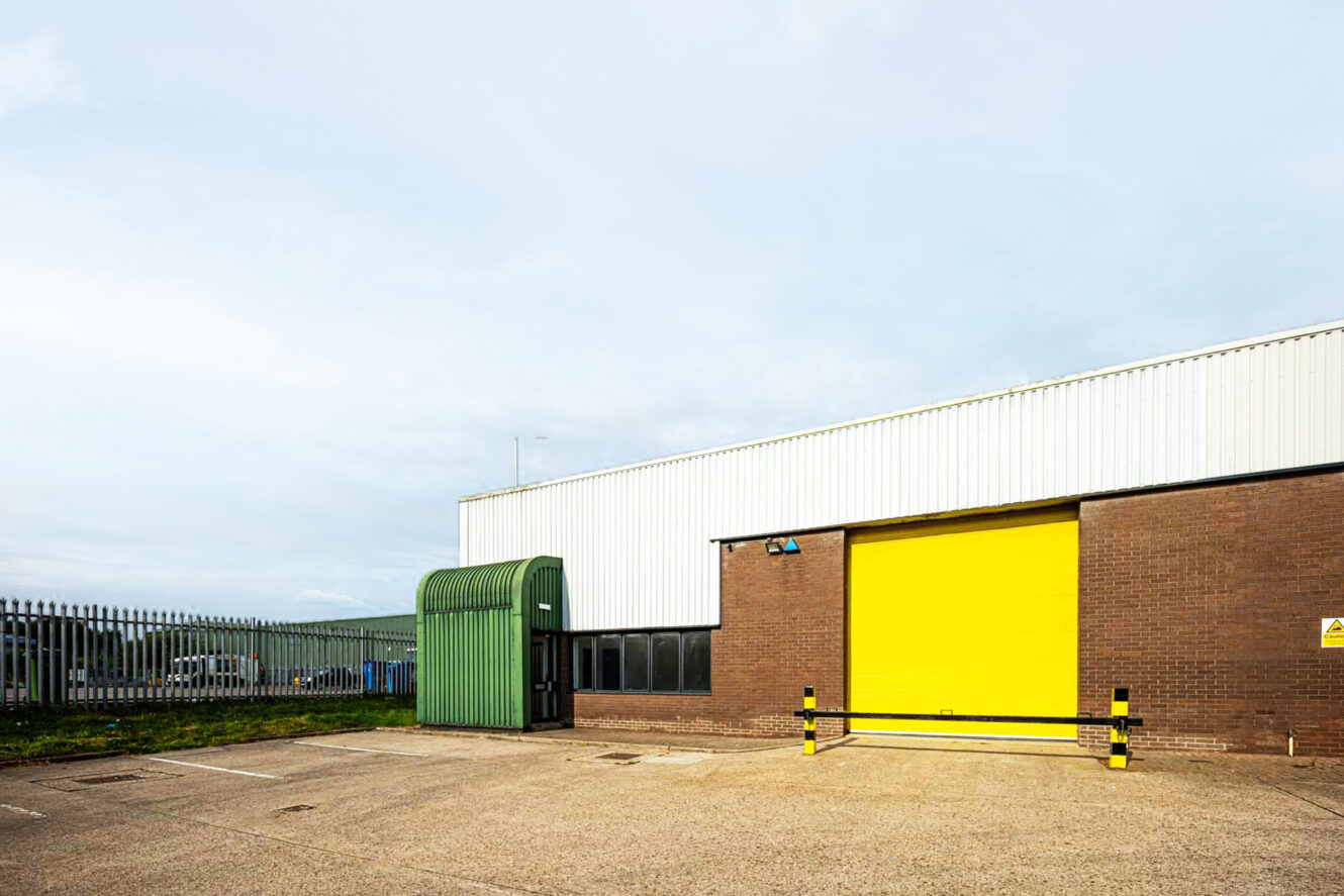 A warehouse with a yellow roller door, brown brick walls, a green metal awning, and an empty paved parking area in front.