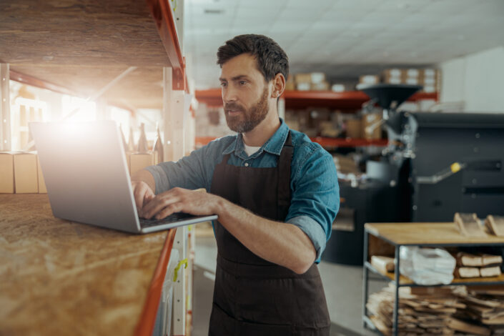 Man wearing an apron stands at a shelf in a warehouse or workshop, working on a laptop with boxes and various supplies visible around him.