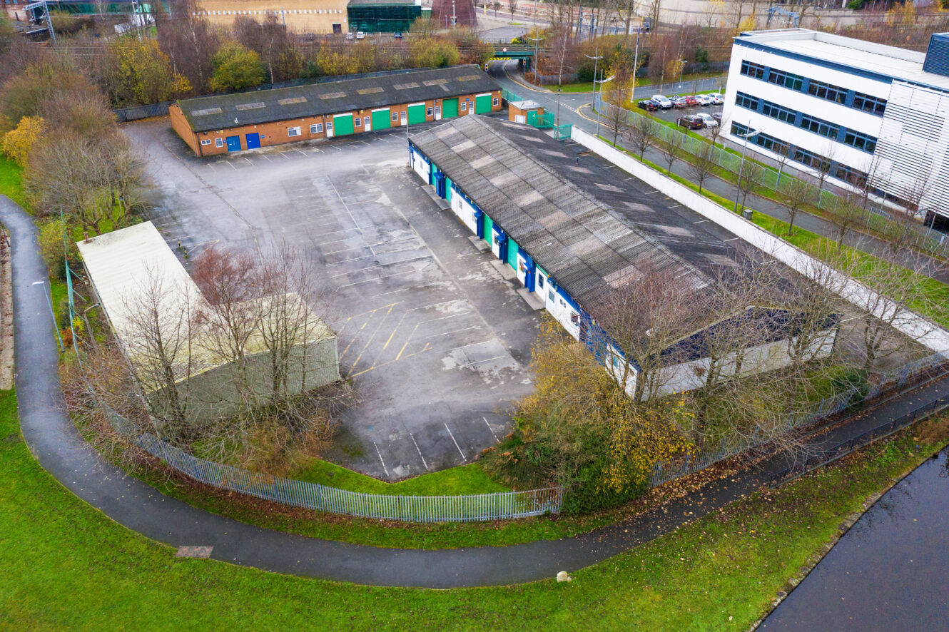 Aerial view of an industrial complex with several warehouses, an empty parking lot, and a nearby office building surrounded by trees and a fence.