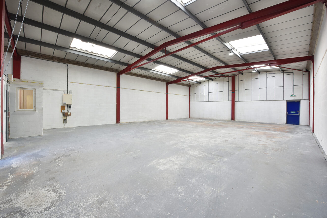 Interior of an empty industrial warehouse with concrete floor, white walls, exposed red steel beams, and a blue door at the far end.
