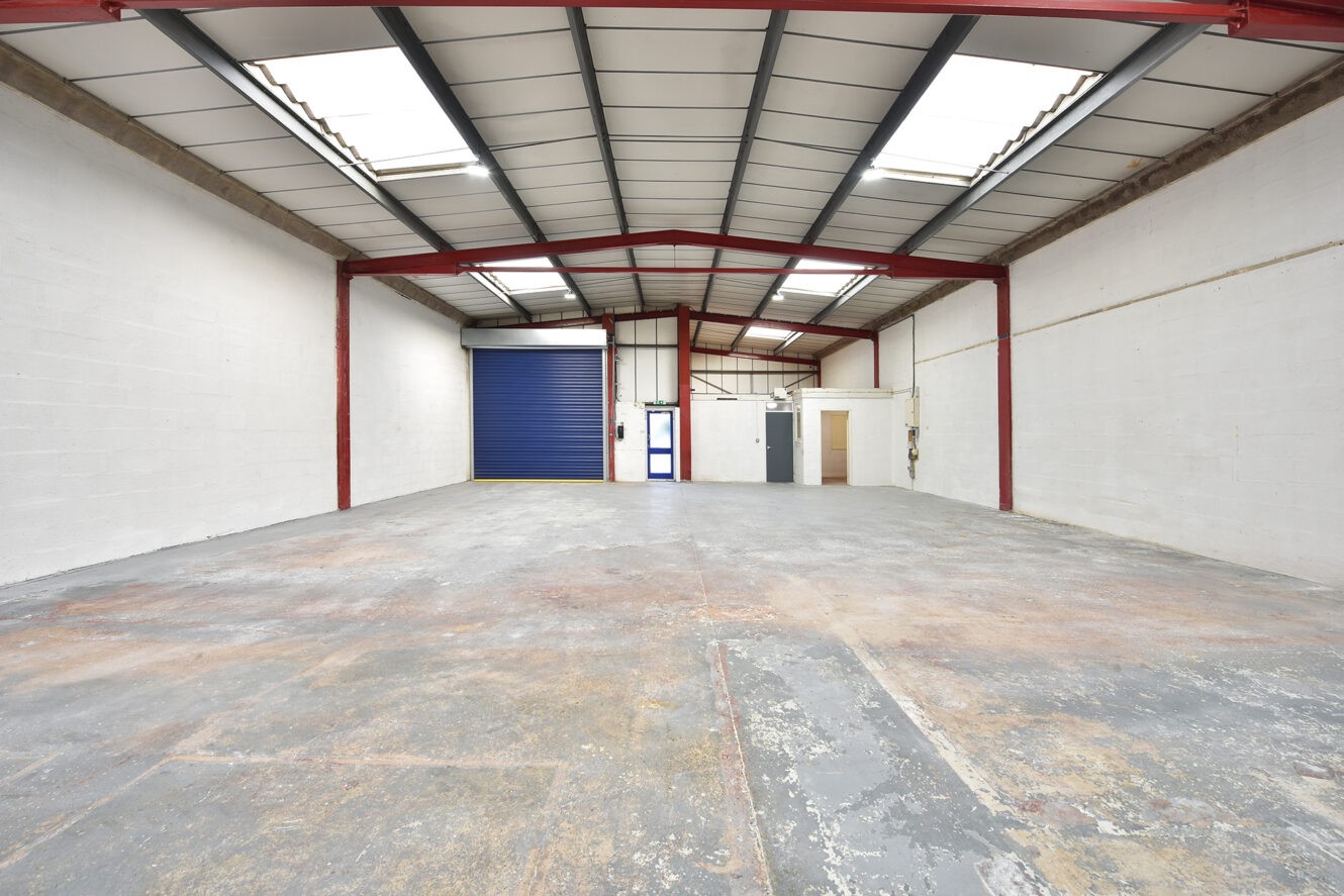 Empty industrial warehouse with high ceiling, exposed beams, skylights, concrete floor, white walls, a blue roller door, and a small enclosed room at the back.