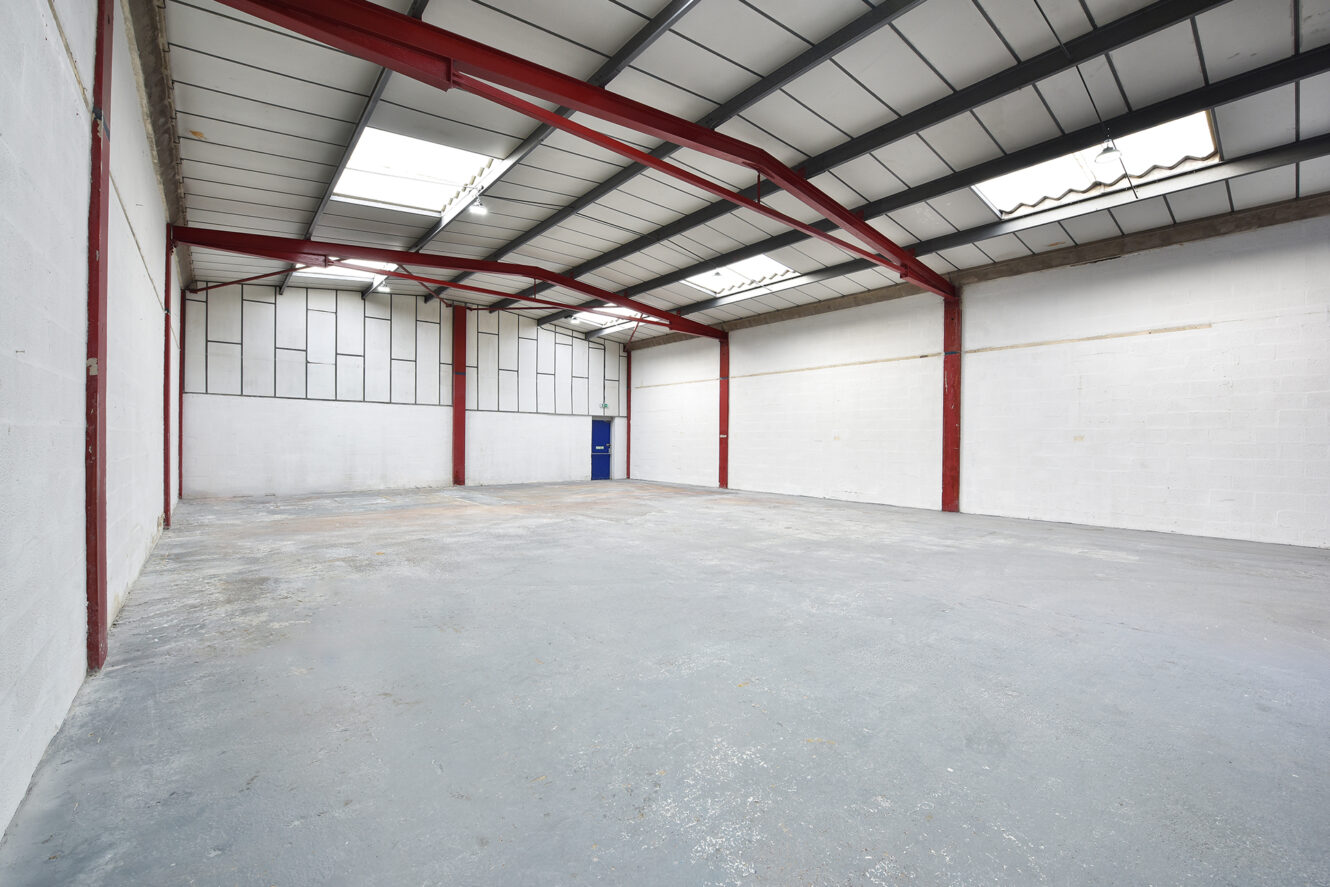 Empty warehouse interior with concrete floor, white brick walls, red steel beams, and skylights in the ceiling; a blue door is visible at the far end.