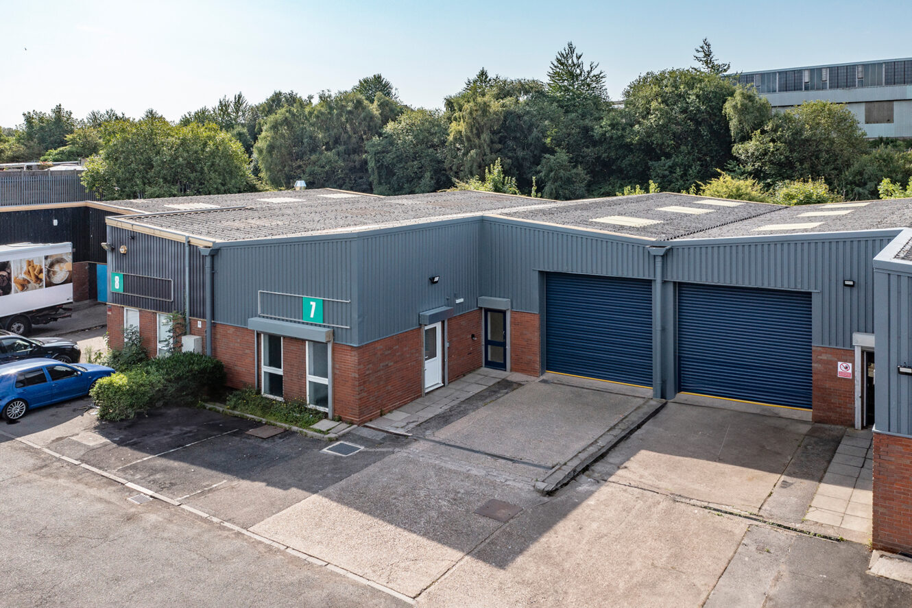 Industrial warehouse unit with two large blue roller doors, brick and gray siding, numbered 7, with parking area and trees in the background.