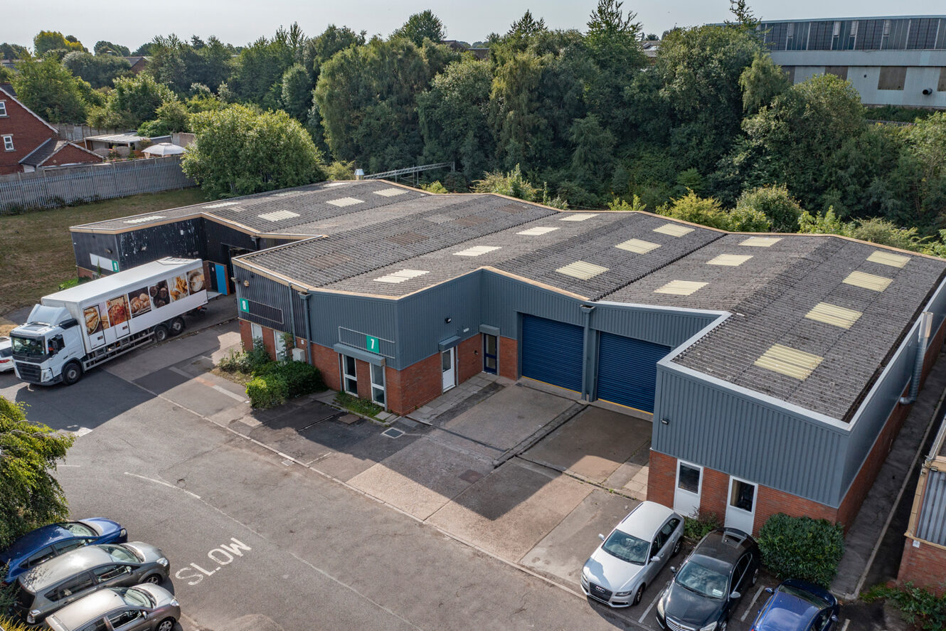 Aerial view of an industrial building with blue doors, cars parked in front, and a delivery truck parked at the side; trees and other buildings in the background.