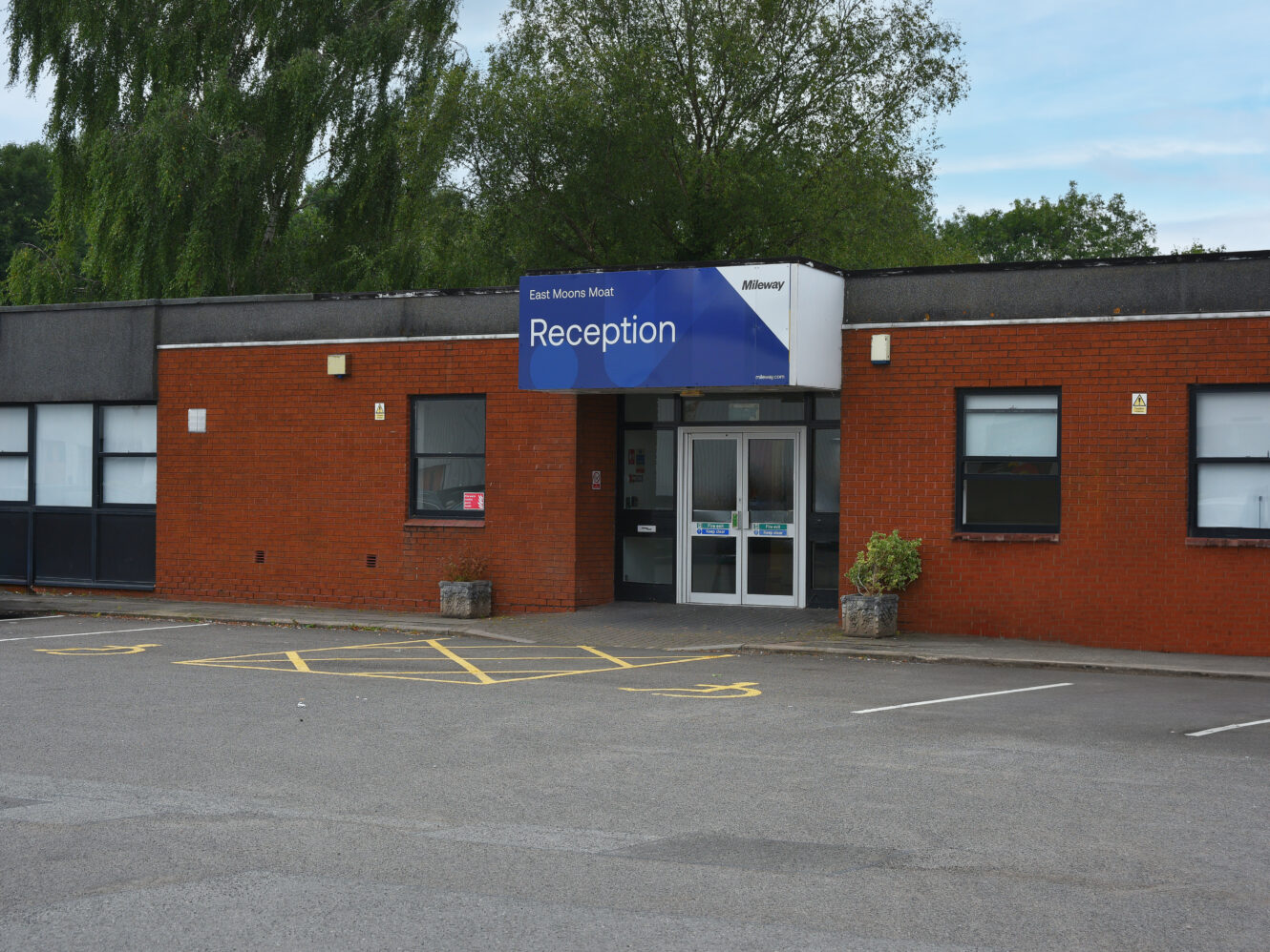 The image shows the entrance to a brick building with a blue sign above the door that reads Reception. There are parking spaces, including a marked accessible spot, in front.