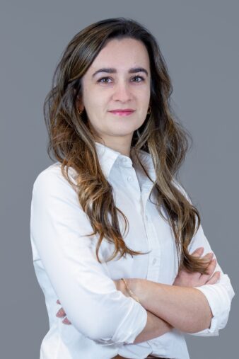 A woman with long brown hair wearing a white button-up shirt stands with arms crossed, facing the camera against a plain gray background.