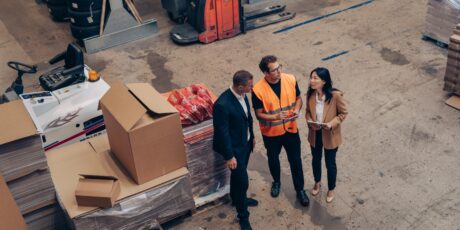 Three people, two in business attire and one in a safety vest, stand in a warehouse discussing near stacks of boxes and pallets.