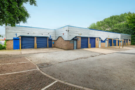 A low industrial building with multiple blue roller shutter doors, numbered 1 to 6, is surrounded by empty parking spaces and trees.