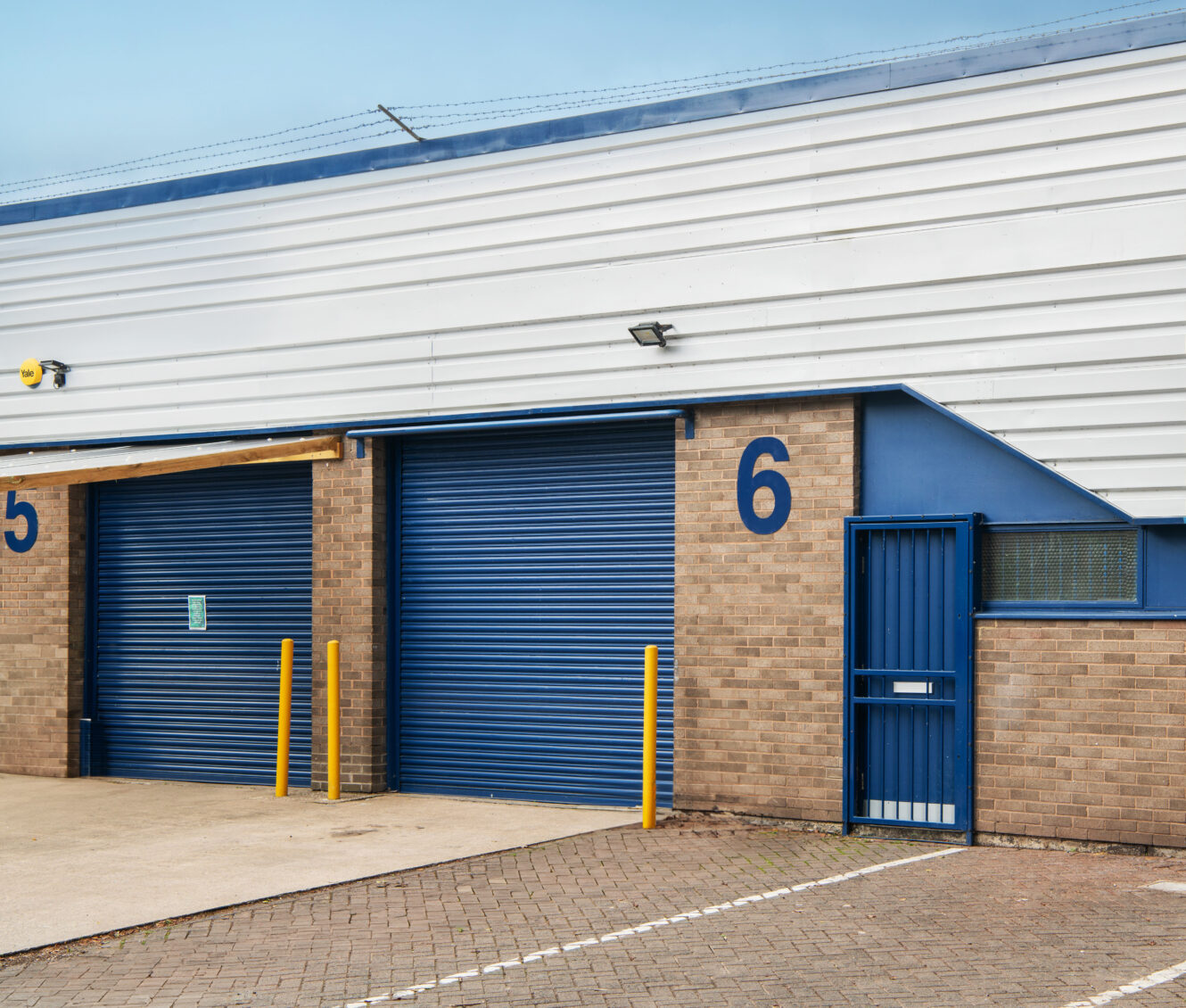 Industrial building exterior with blue roller shutter doors, marked with numbers 5 and 6, yellow bollards, and a blue metal entry gate.