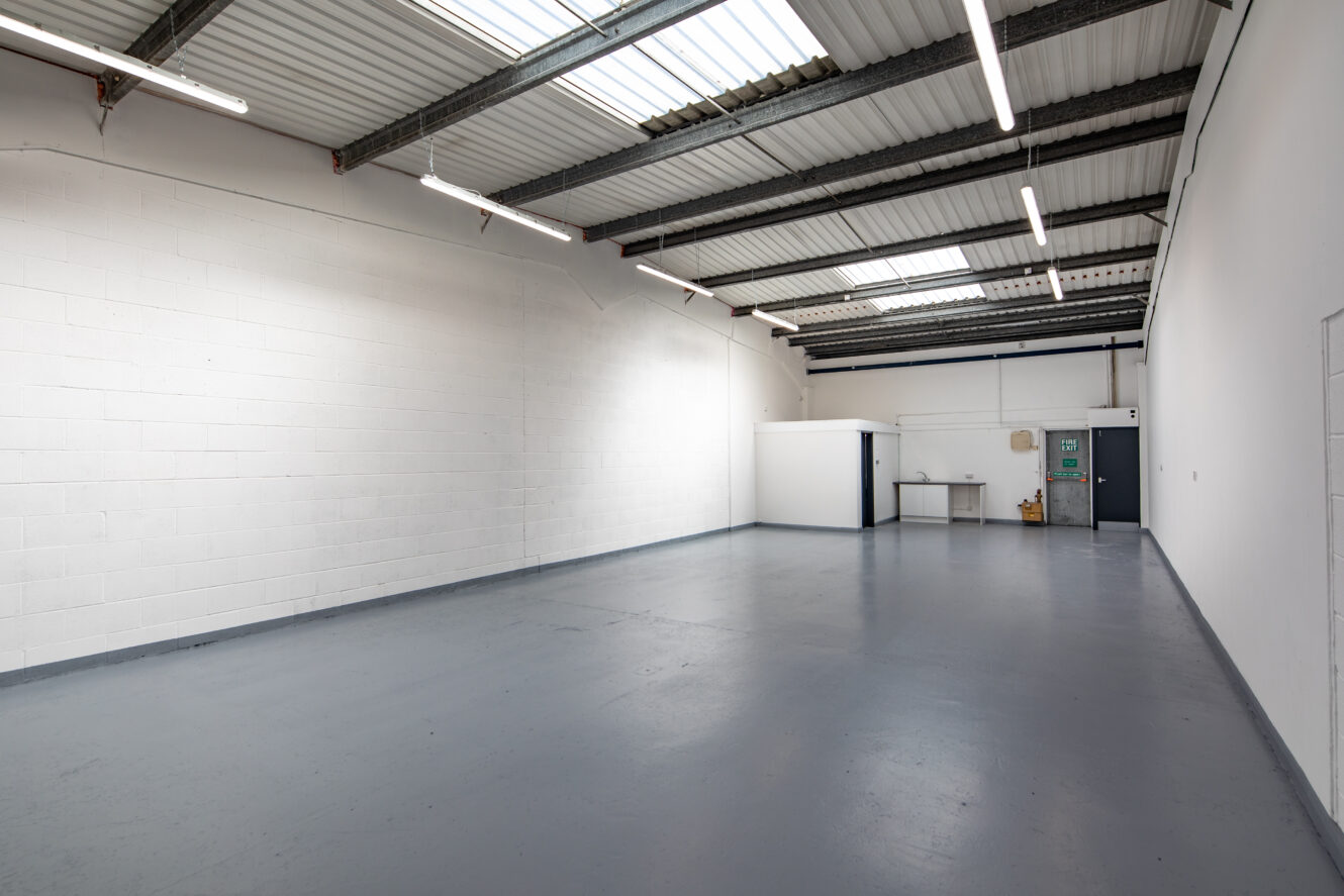 Empty industrial warehouse with white walls, grey concrete floor, high ceiling, exposed beams, and fluorescent lighting. A small office area and utility panel are visible at the back.