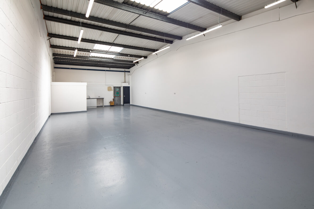 Empty industrial warehouse with white walls, grey floor, exposed ceiling beams, fluorescent lighting, and a small utility area in the back corner.