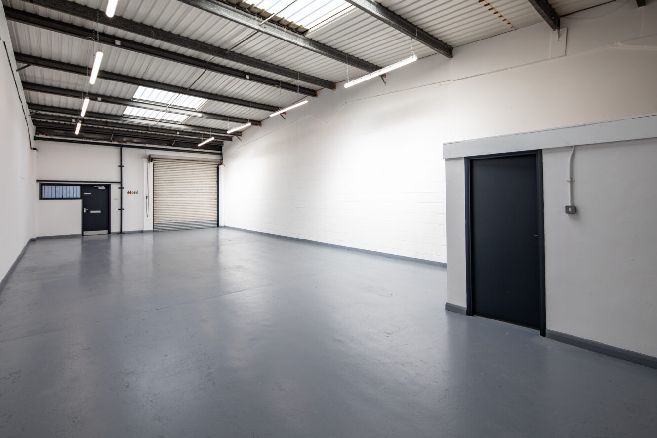 Empty industrial warehouse unit with gray floor, white walls, high ceiling, fluorescent lights, roller shutter door, and two black doors.