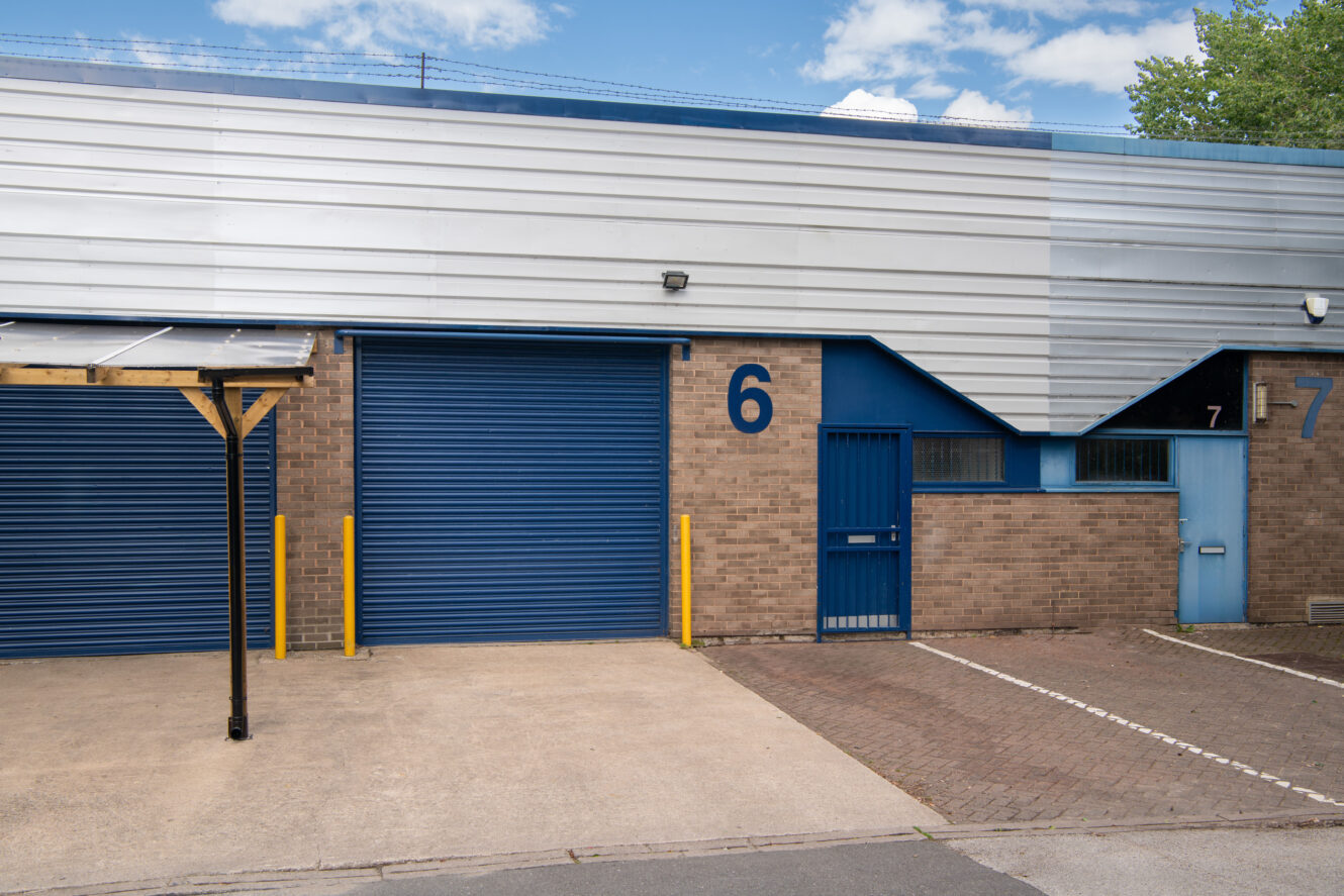 A commercial unit with brown brick walls, blue roller shutter door, blue entry door, and the number 6 displayed next to the door. A parking area is in front.