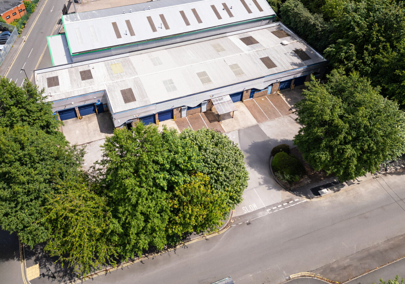 Aerial view of a large industrial warehouse with blue loading bays, surrounded by trees and adjacent to a road marked with SLOW.
