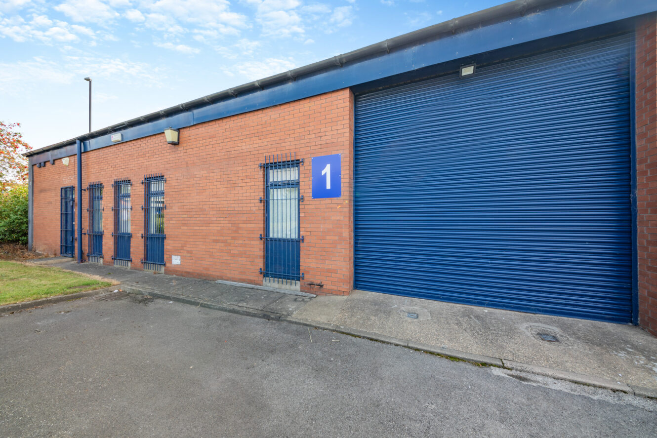 Single-story brick industrial unit with blue doors, barred windows, and a large blue roller shutter numbered 1 on the wall.