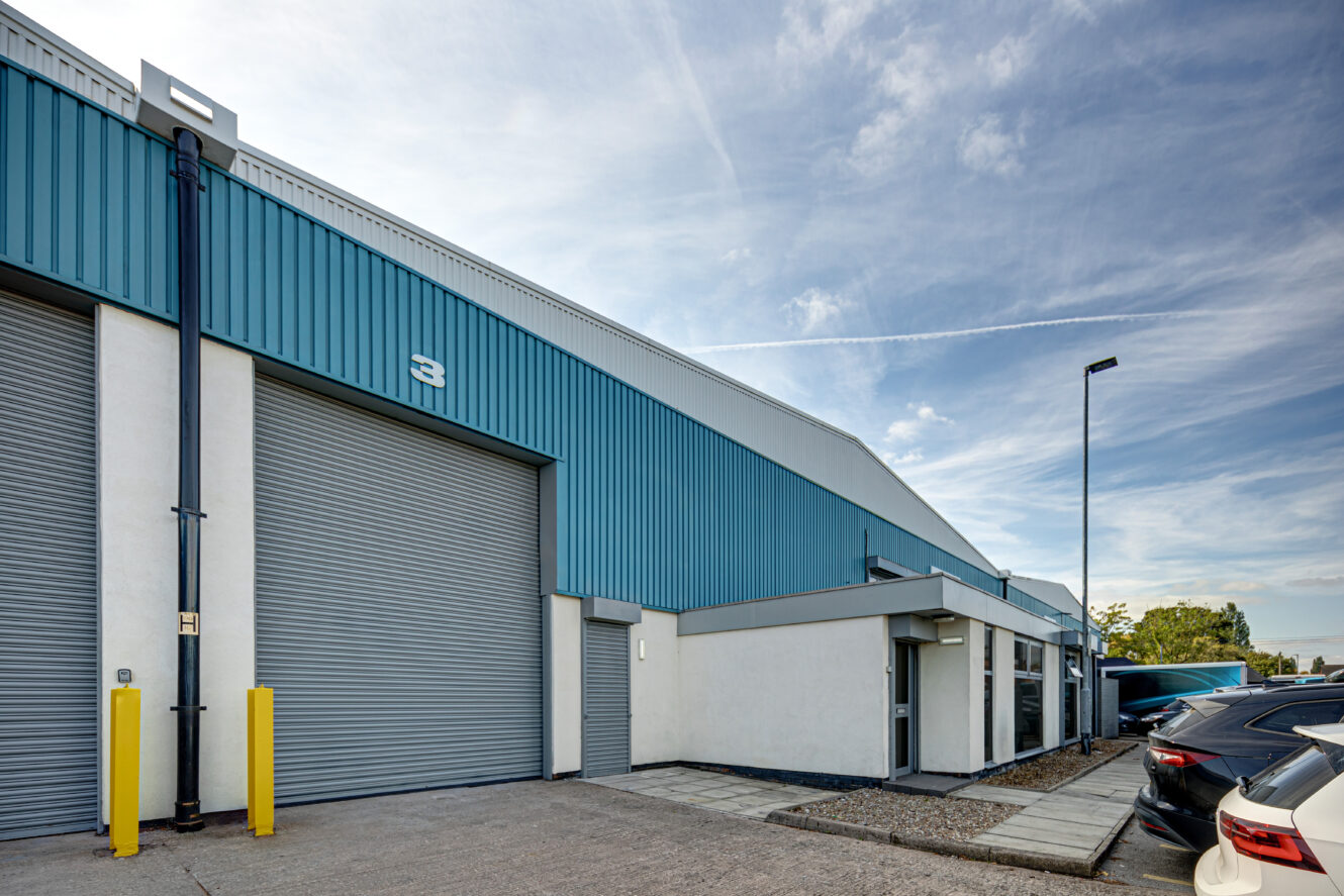 Industrial warehouse building with blue and white exterior, large roller shutter doors, parked cars, and clear sky in the background.