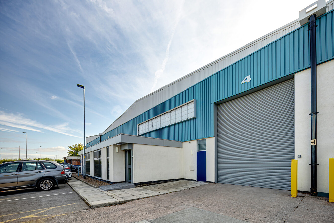 Exterior view of a modern industrial warehouse with a blue and white facade, large roller shutter door, and parked cars on the left.