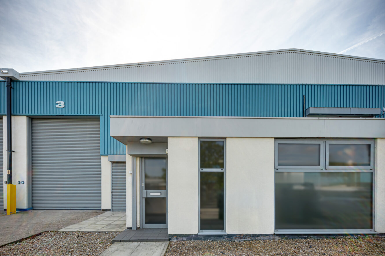 Exterior view of an industrial warehouse building with blue metal siding, large garage door labeled 3, and adjacent office entrance with windows.