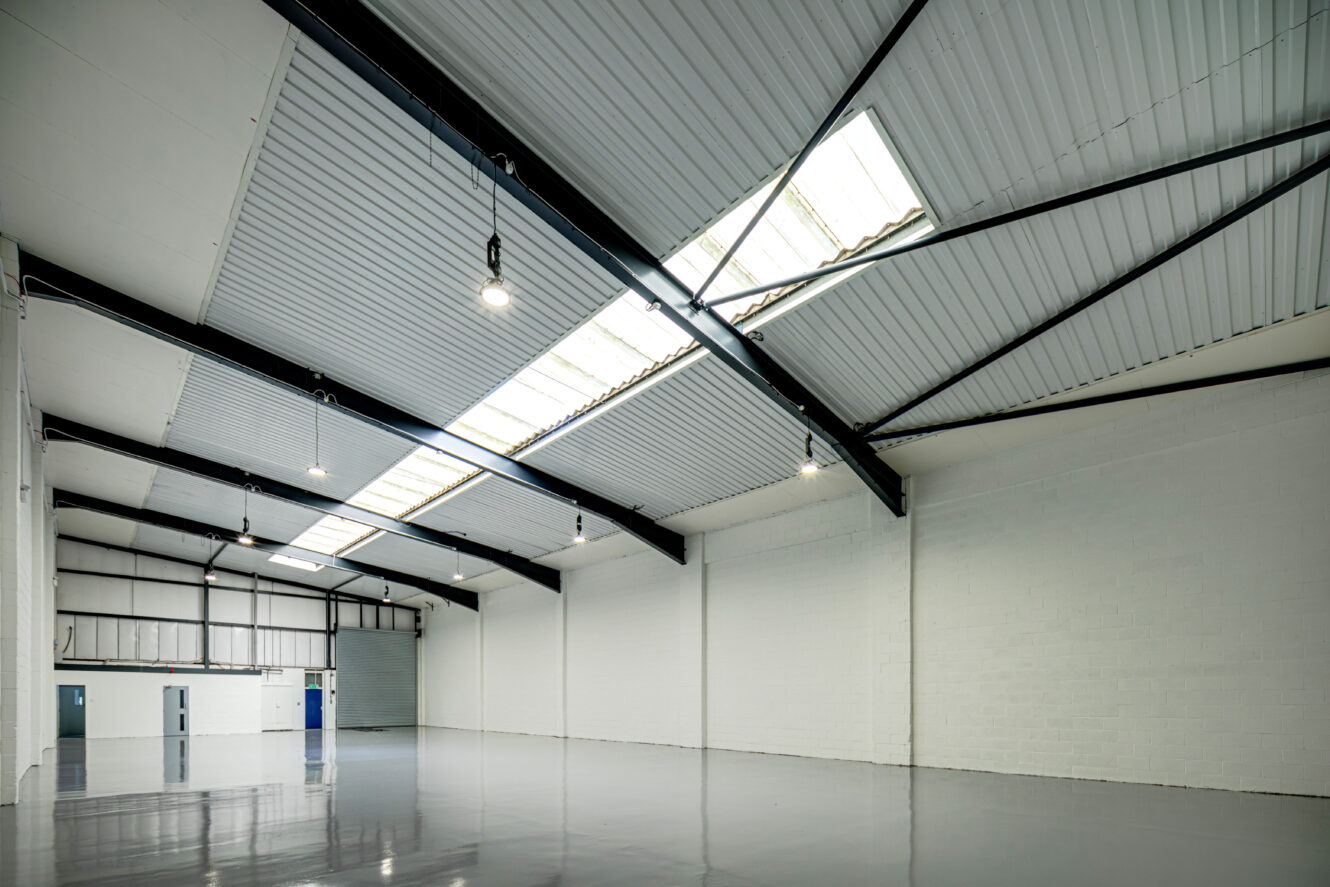 Large, empty industrial warehouse with white walls, high vaulted ceiling, exposed beams, skylights, and polished gray concrete floor.