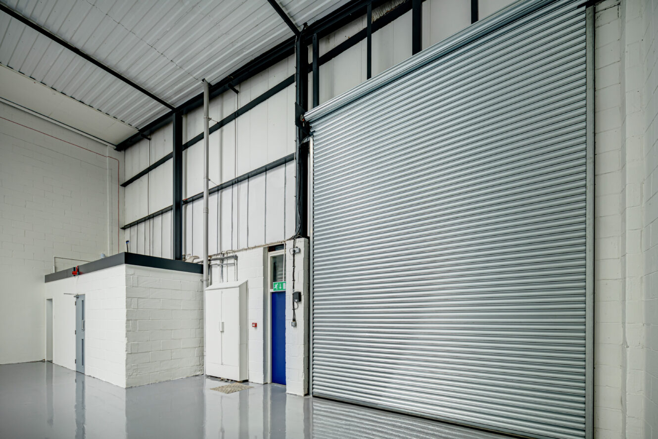 Interior of an empty industrial warehouse with a large metal roller shutter door, white brick walls, a small enclosed office, and polished gray floor.