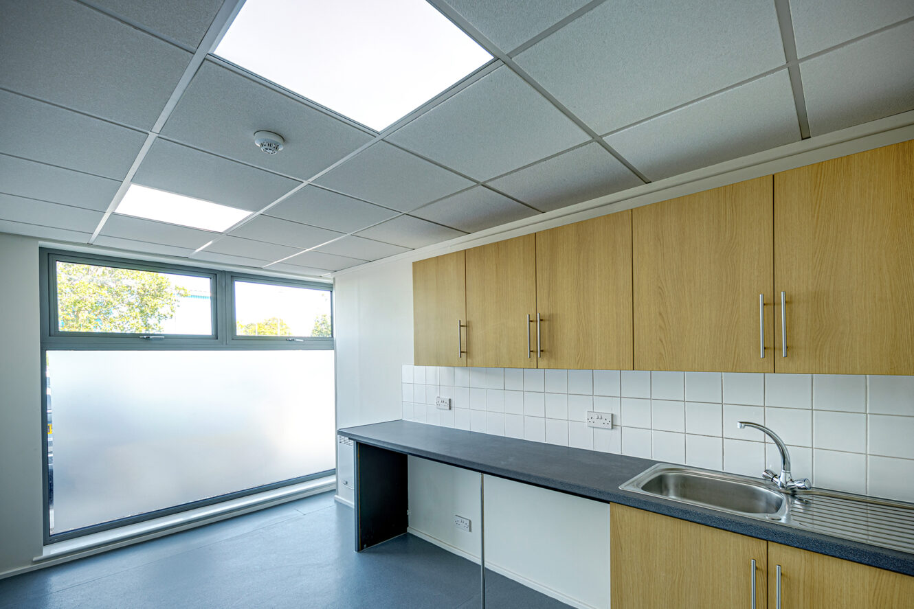 Modern kitchenette with wood cabinets, grey countertop, stainless steel sink, tiled backsplash, and large frosted window letting in natural light.
