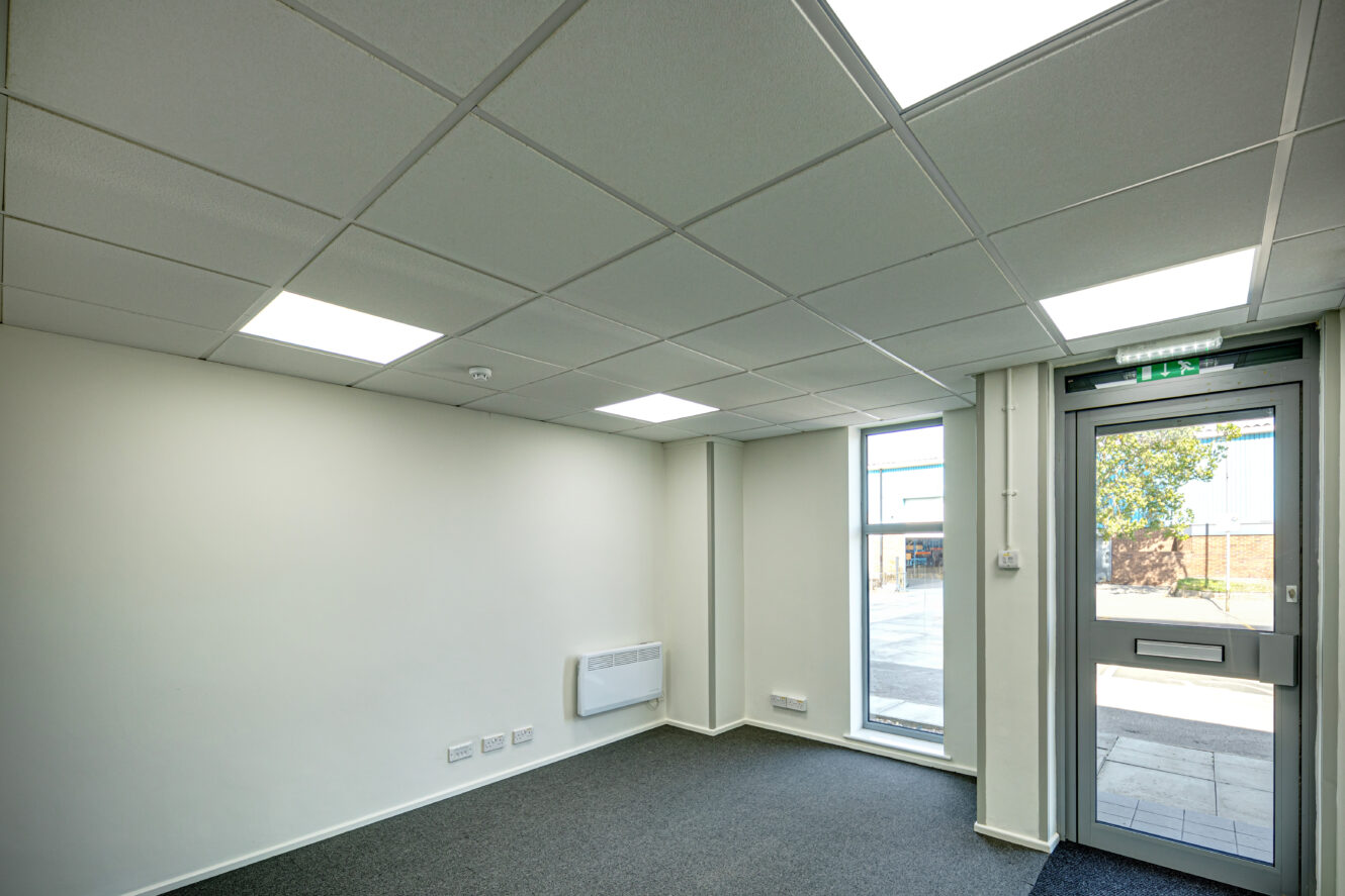 Empty office room with white walls, gray carpet, a window, a door with a mail slot, overhead fluorescent lights, and an electric wall heater.