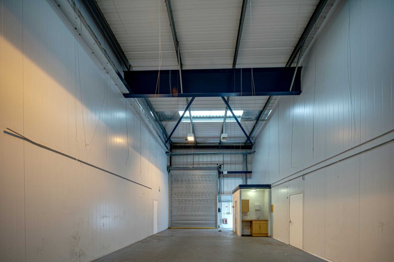 Interior of an empty industrial warehouse with white walls, a high ceiling, a roller shutter door, overhead beams, and a small office area to the right.