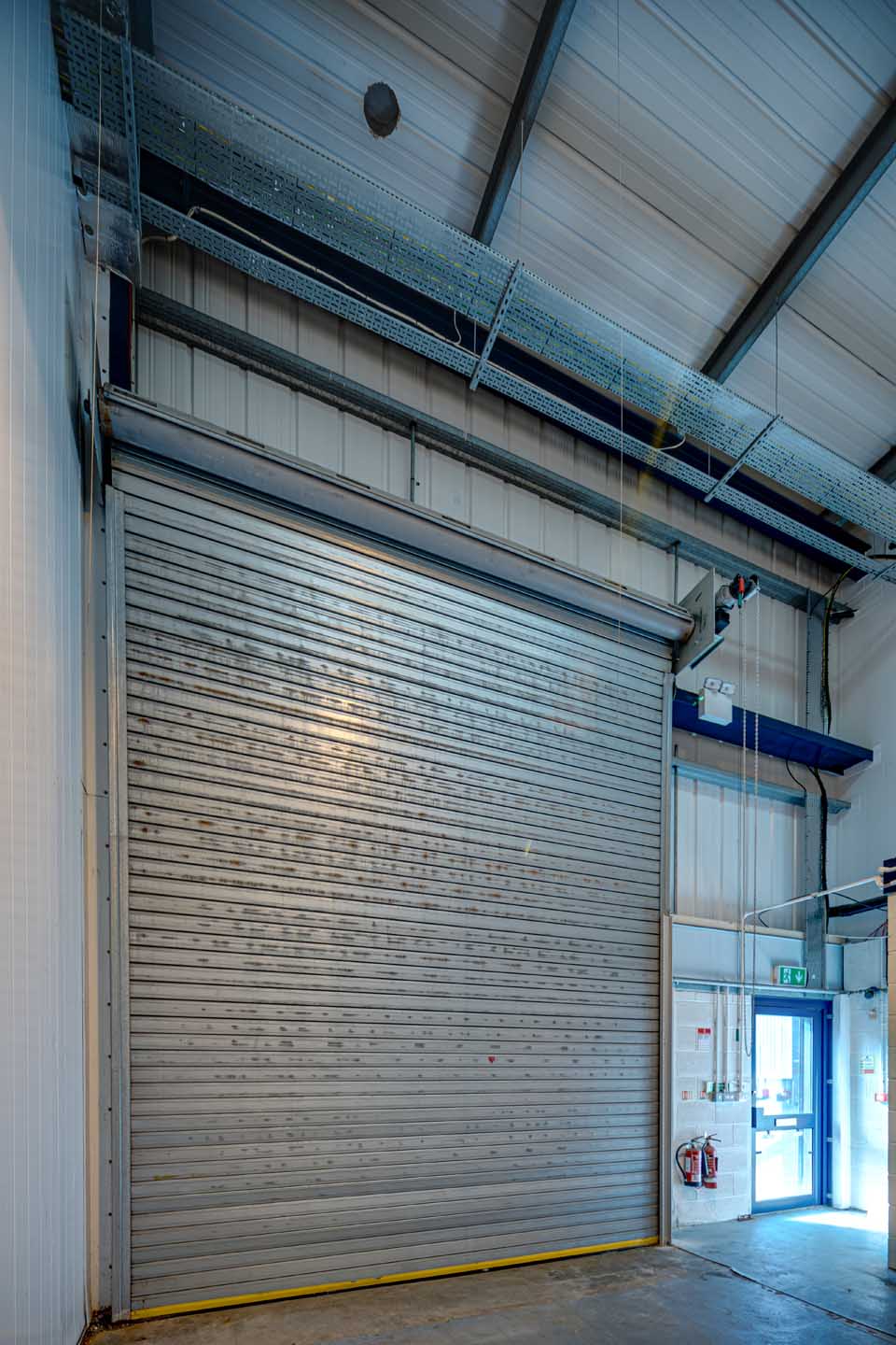 Large metal roller shutter door in an industrial warehouse, with a small exit door and fire extinguishers on the right side.