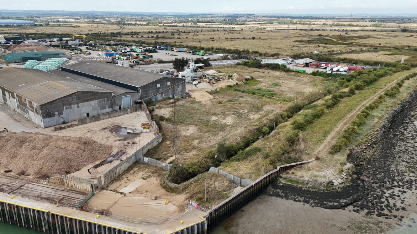 Aerial view of an industrial waterfront area with warehouses, storage piles, vehicles, and surrounding open land next to a river or shoreline.