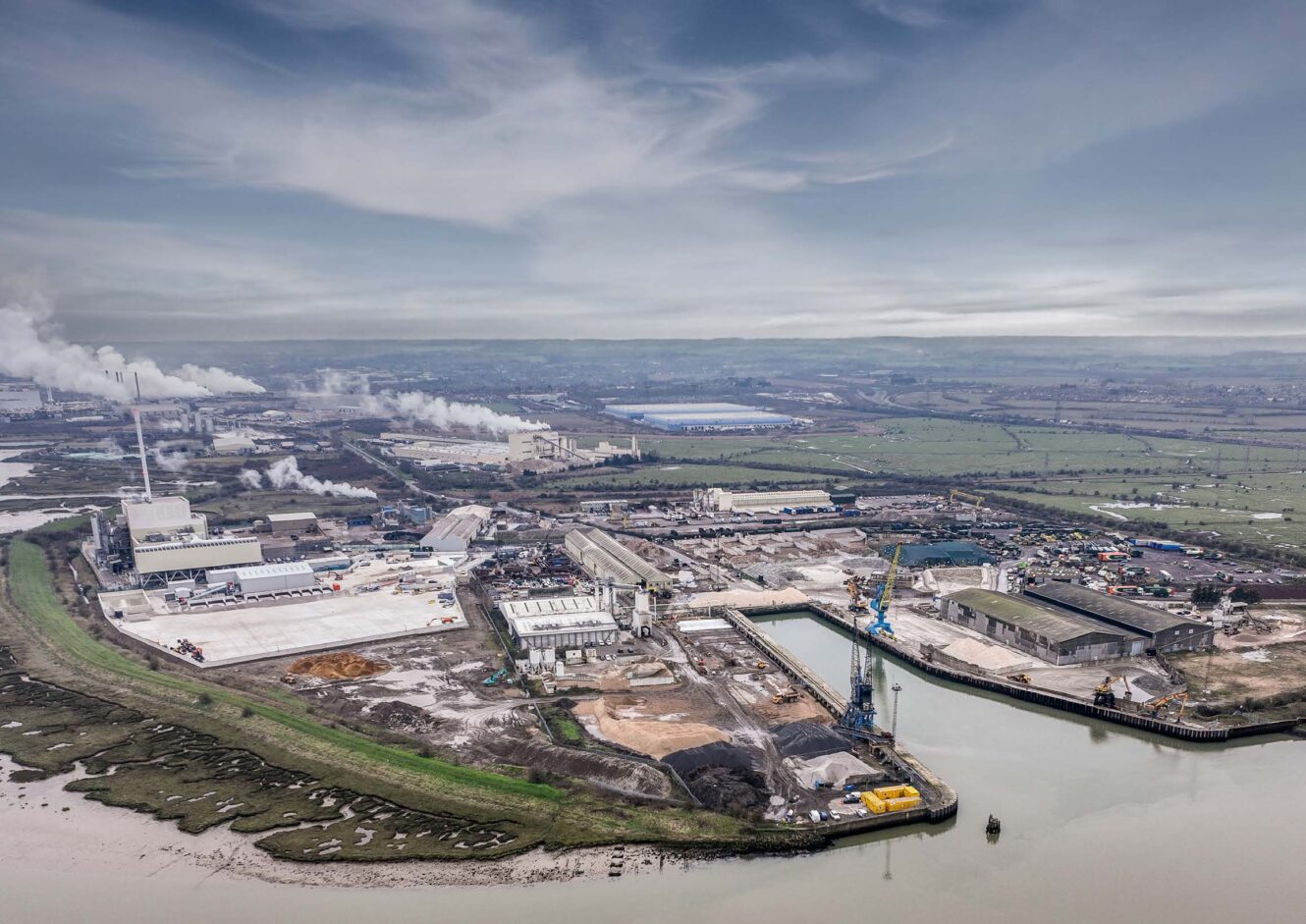 Aerial view of an industrial complex with factories, warehouses, smoke stacks, and a dock alongside a river, set in a rural landscape.