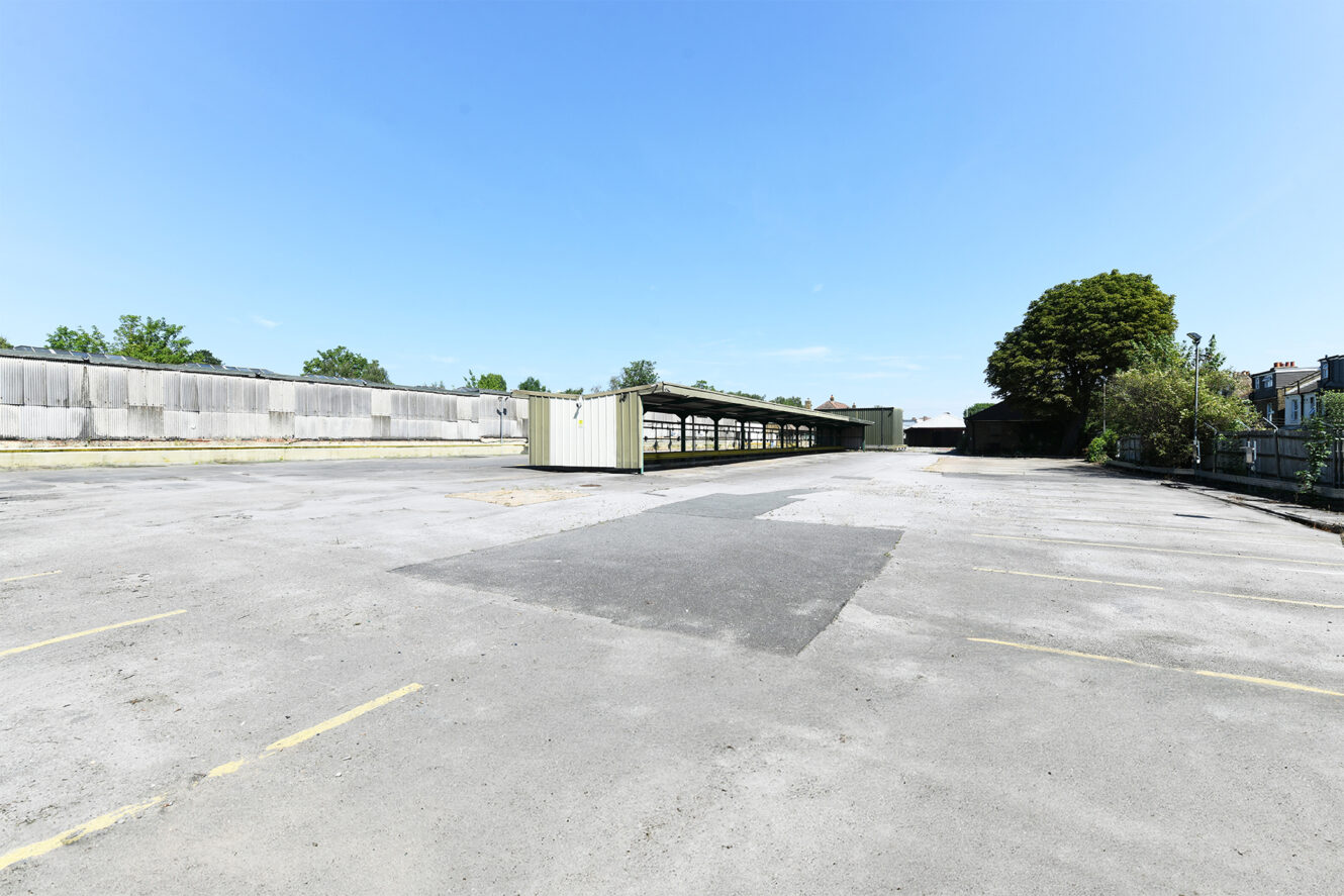 An empty, expansive parking lot with yellow lines, a few small sheds, and trees and buildings in the background under a clear blue sky.