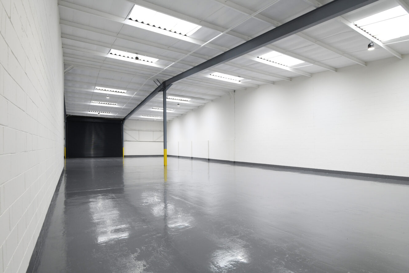 Interior of a large, empty warehouse with white walls, grey polished floor, skylights, and a black roller shutter door at the far end.