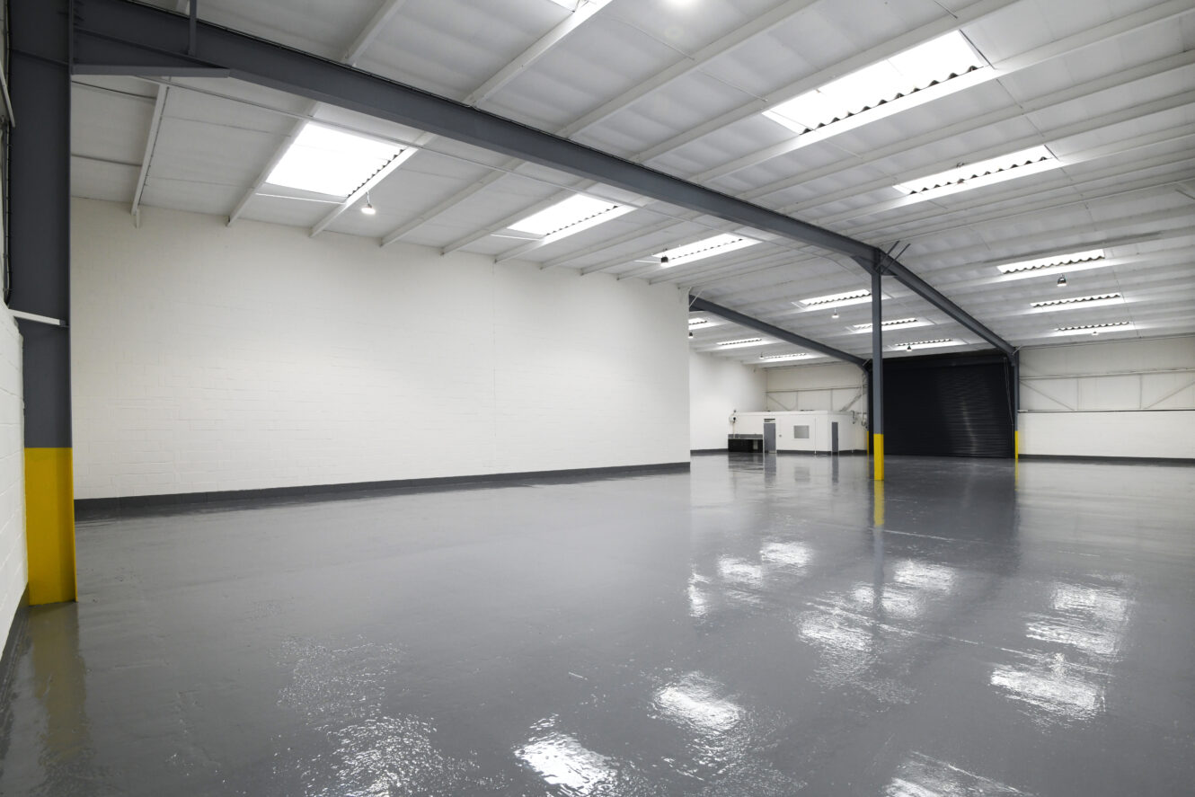 Interior of an empty industrial warehouse with white walls, grey floors, exposed beams, and overhead lighting.