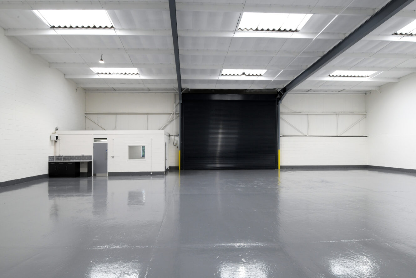 An empty industrial warehouse with white walls, grey polished floor, a small office area on the left, and a large black roller shutter door at the back.