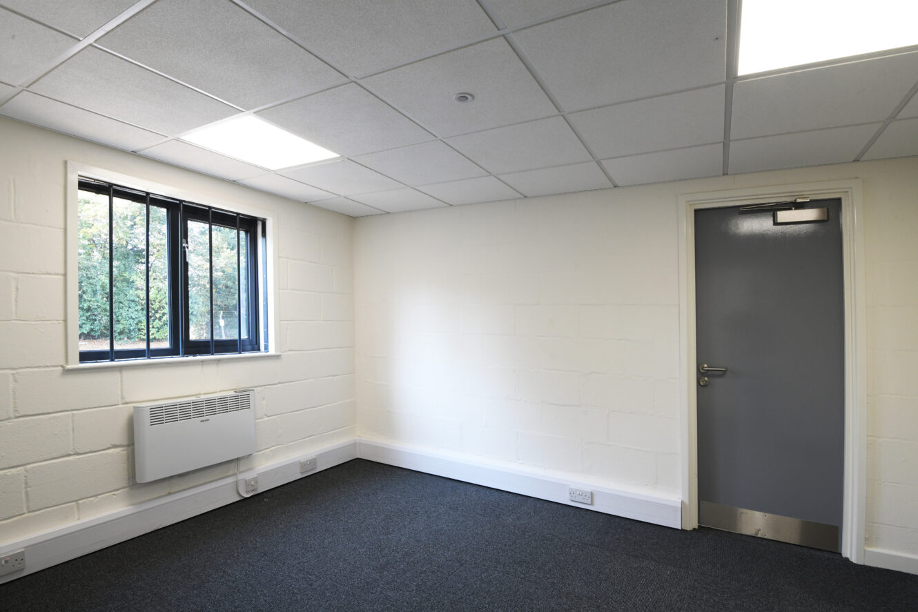 Empty room with white painted brick walls, a gray door, barred window, heater, and blue-gray carpeted floor under fluorescent ceiling lights.