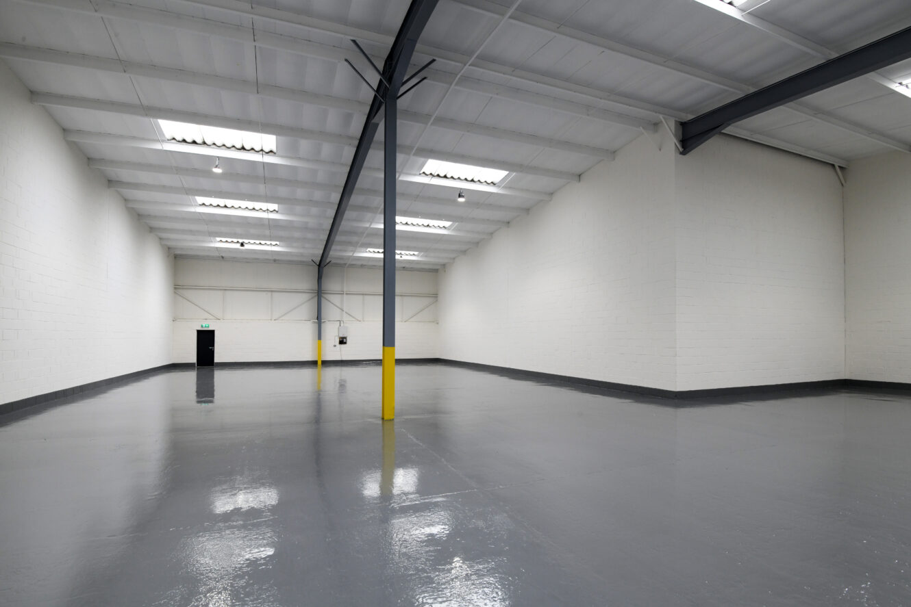 Large, empty warehouse with white walls, gray polished floor, exposed beams, fluorescent lighting, and support columns, one of which is painted yellow at the base.