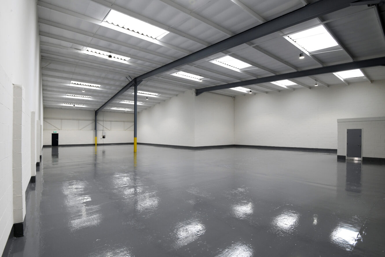 Empty warehouse with white walls, gray glossy floor, high ceiling, exposed beams, skylights, and two doors on opposite sides.