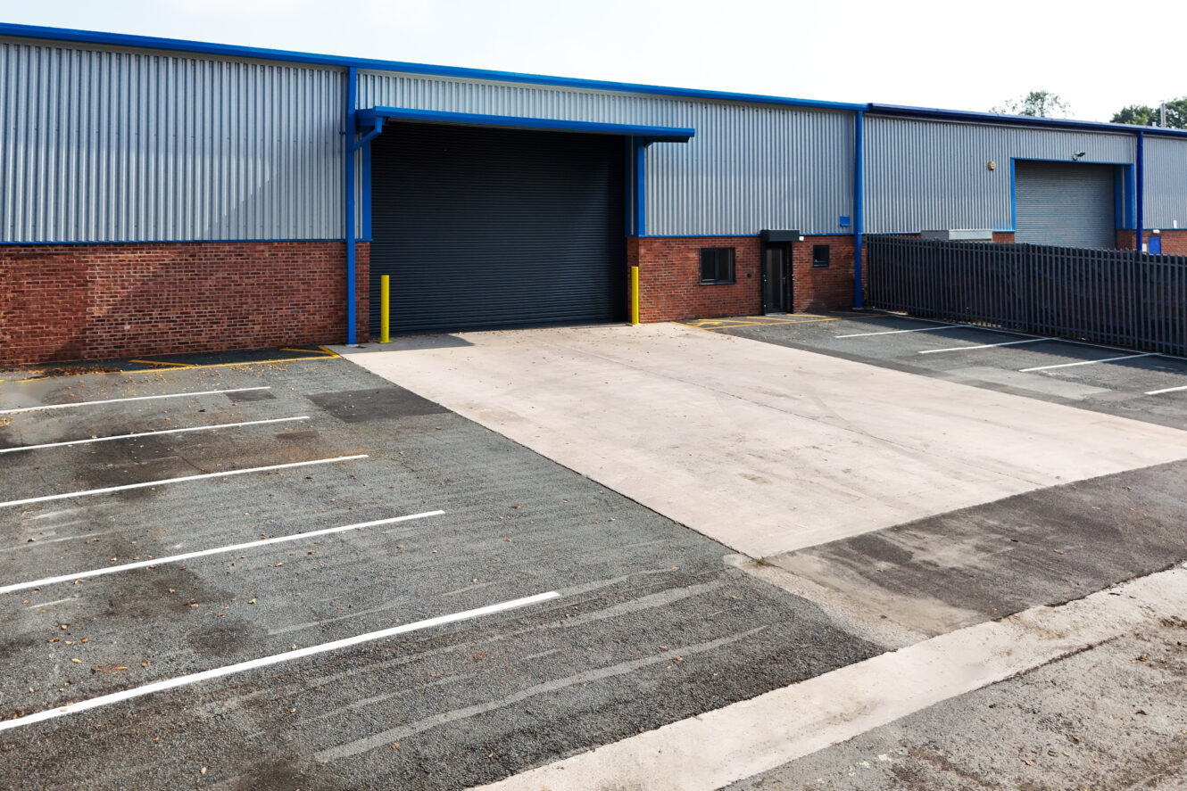A large industrial warehouse with a brick and metal exterior, a closed black roller door, and an empty parking lot with marked spaces in front.