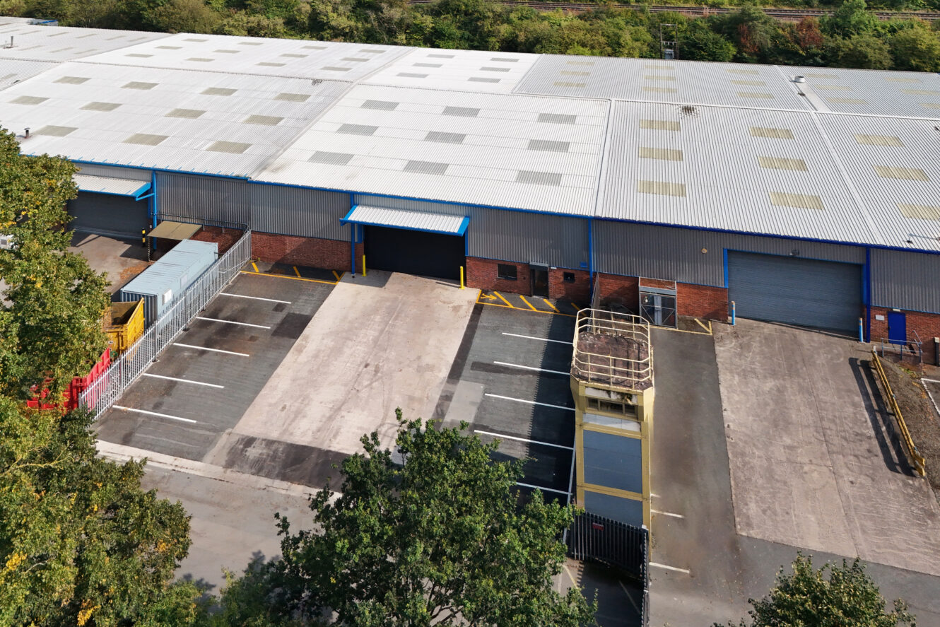 Aerial view of an industrial warehouse with loading bays, empty parking spaces, a fenced area, and adjacent trees.