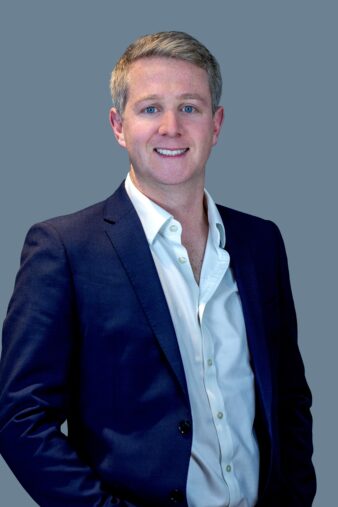 A man in a dark blue suit and white shirt stands against a plain blue background, smiling at the camera.